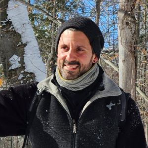 A man wearing a black beanie, scarf, and black jacket stands outdoors in a snowy forest, smiling with sunlight on his face during a guided snowshoeing tour. A man wearing a black beanie, scarf, and black jacket stands outdoors in a snowy forest, smiling with sunlight on his face during a guided snowshoeing tour.
