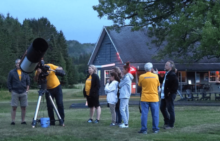 A group of people stand on grass near a large telescope, stargazing and exploring astronomy; some look through the telescope while others observe. A building and trees are in the background, suggesting an evening gathering.