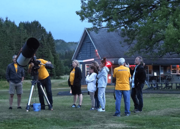 A group of people stand on grass near a large telescope, stargazing and exploring astronomy; some look through the telescope while others observe. A building and trees are in the background, suggesting an evening gathering.