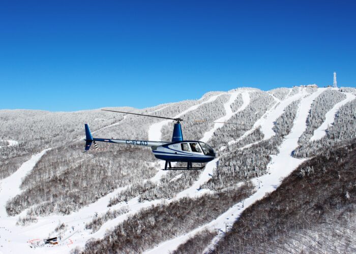 A blue and white helicopter flies over a snow-covered mountain with ski slopes, offering breathtaking scenic flights under a clear blue sky.