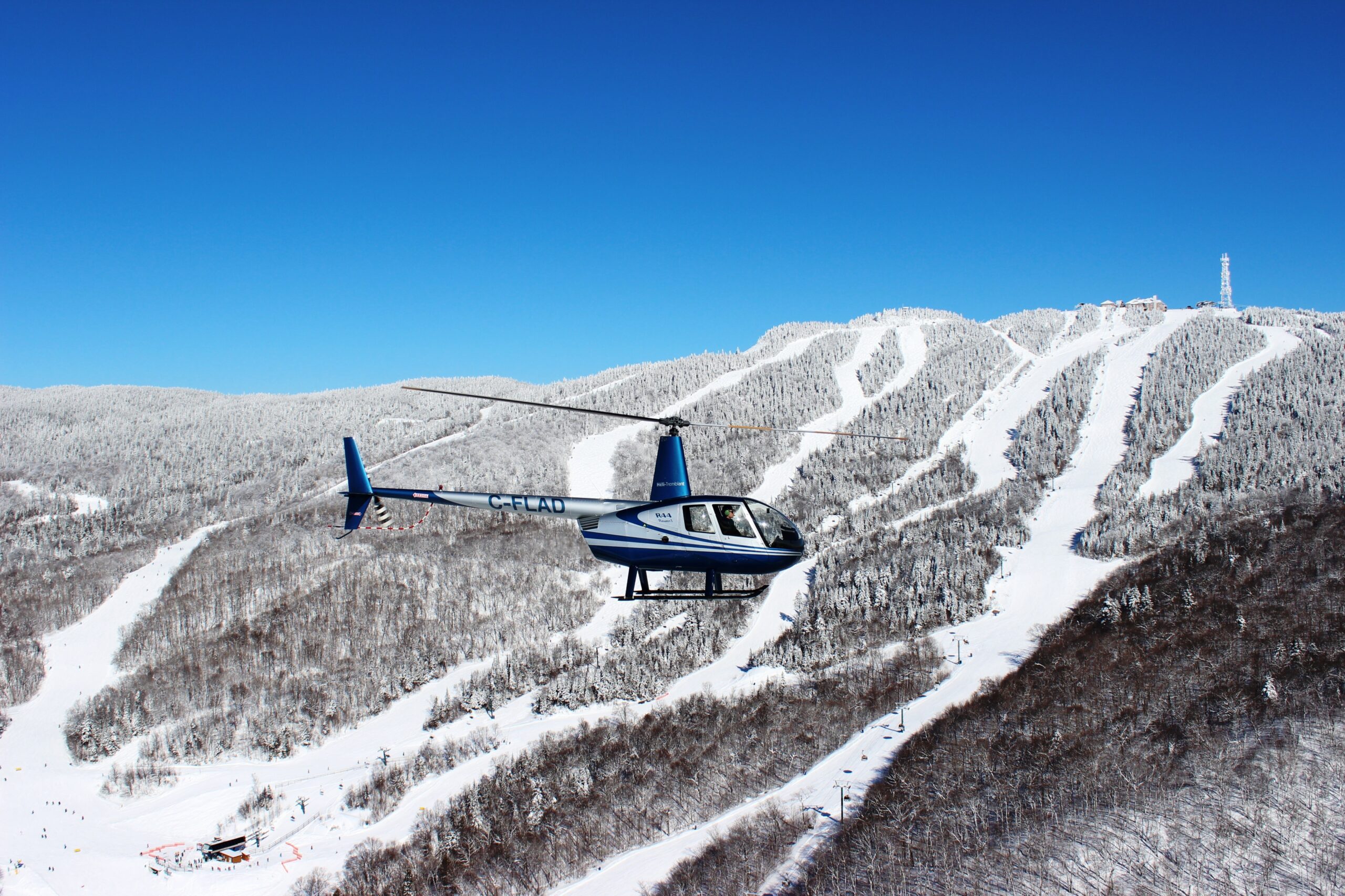 A blue and white helicopter flies over a snow-covered mountain with ski slopes, offering breathtaking scenic flights under a clear blue sky.