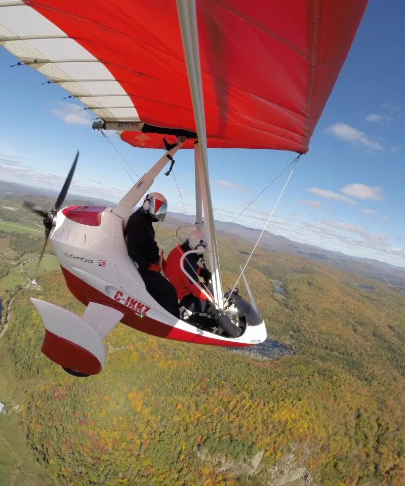 Une personne s'initie au vol ultraléger en pilotant un autogire rouge et blanc (ULM) qui survole une mosaïque de champs, de collines et de forêts sous un ciel bleu parsemé de nuages.