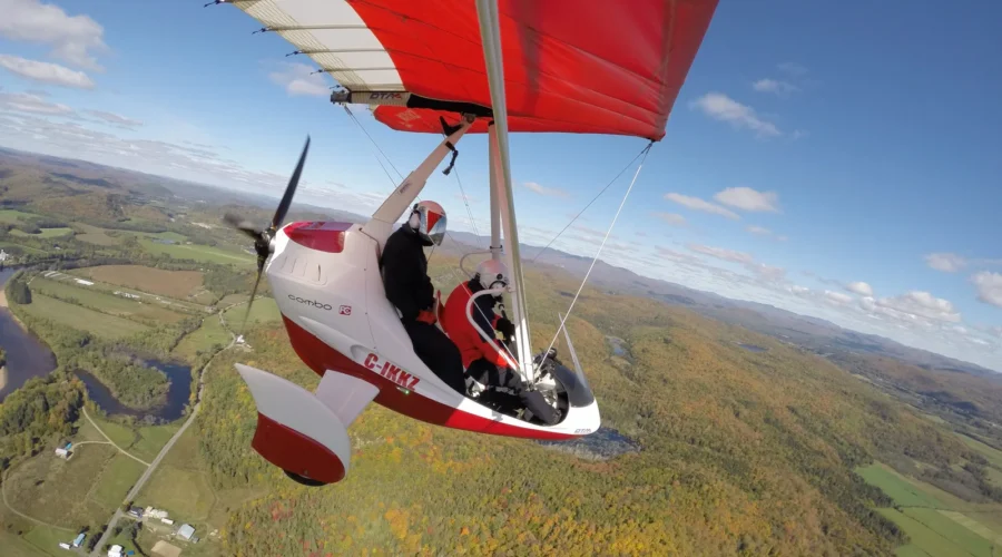 A red and white ultralight flight aircraft with two people onboard flies above a landscape of fields, forest, and hills under a blue sky with scattered clouds. A red and white ultralight flight aircraft with two people onboard flies above a landscape of fields, forest, and hills under a blue sky with scattered clouds.