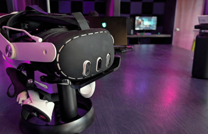 A VR headset and controllers rest on a stand on a blue table, hinting at an immersive Arcade VR experience; a water bottle and computer setup are visible in the background, perfect for those considering headset rental.