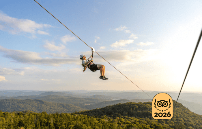 Person ziplining over the forested mountain landscape of Mont-Tremblant under a blue sky, with a "2026" badge in the lower right corner.