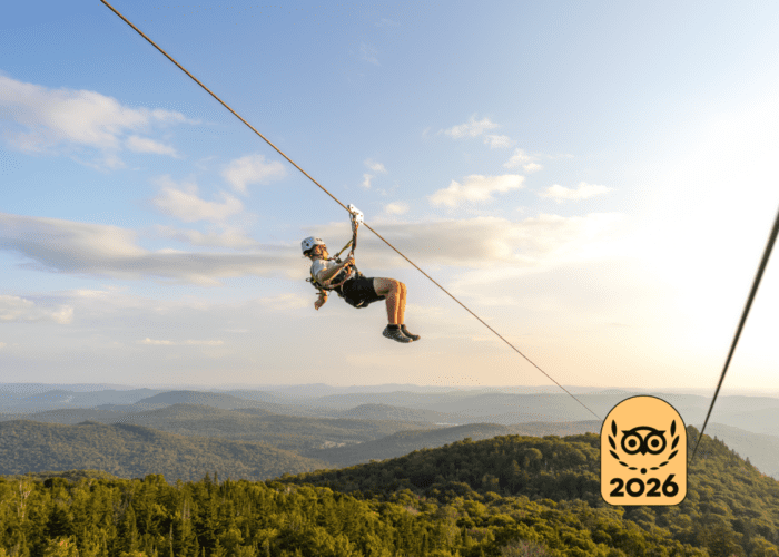 Person ziplining over the forested mountain landscape of Mont-Tremblant under a blue sky, with a "2026" badge in the lower right corner.