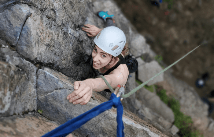 Une personne portant un casque escalade une paroi rocheuse à la Montagne d'Argent, s'étirant vers le haut tout en étant attachée à une corde lors d'une aventure d'escalade.