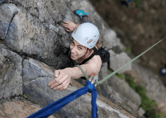 A person wearing a white helmet and climbing gear ascends the steep rock face of Montagne d'Argent, reaching up toward the camera while secured by a rope during a thrilling rock-climbing adventure.