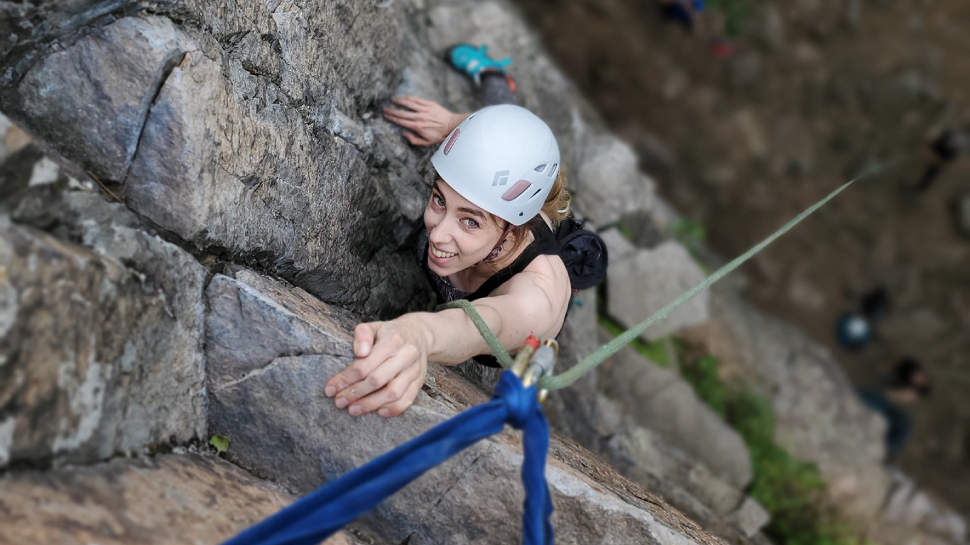 A person wearing a white helmet and climbing gear ascends the steep rock face of Montagne d'Argent, reaching up toward the camera while secured by a rope during a thrilling rock-climbing adventure.