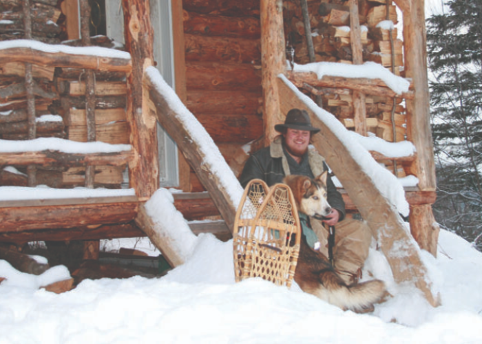 A seasoned woodsman wearing winter clothing sits with a dog beside wooden snowshoes on the snowy porch of a log cabin, showcasing his outdoor skills.