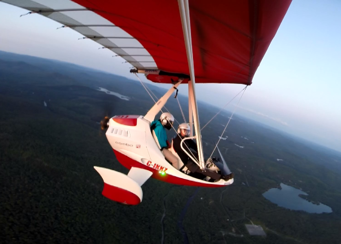 Two people wearing helmets experience ultralight flight as they pilot a small, red-and-white open-cockpit aircraft high above a landscape with forests, lakes, and rivers.