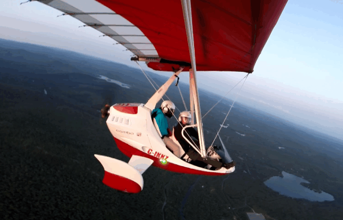 Two people wearing helmets take flight initiation in a small red and white ultralight airplane, soaring over a green landscape with lakes visible below. Two people wearing helmets take flight initiation in a small red and white ultralight airplane, soaring over a green landscape with lakes visible below.