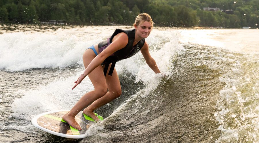 A person wearing a life jacket and swimsuit enjoys wakesurf watersports, riding a wave on a surfboard in a lake with green trees in the background. A person wearing a life jacket and swimsuit enjoys wakesurf watersports, riding a wave on a surfboard in a lake with green trees in the background.