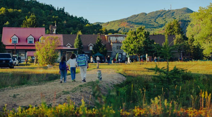 Deux personnes avec des sacs à dos marchent sur un sentier vers un pavillon entouré d'arbres dans le parc du Domaine Saint-Bernard, avec les montagnes visibles en arrière-plan - parfait pour une excursion guidée ou une observation d'oiseaux.