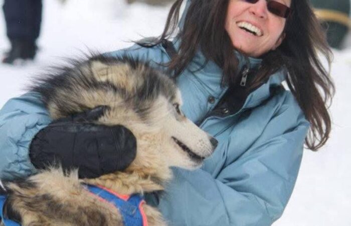 Marie-Claude, in a blue winter jacket, smiles warmly as she holds a large, furry dog outdoors in a snowy setting.