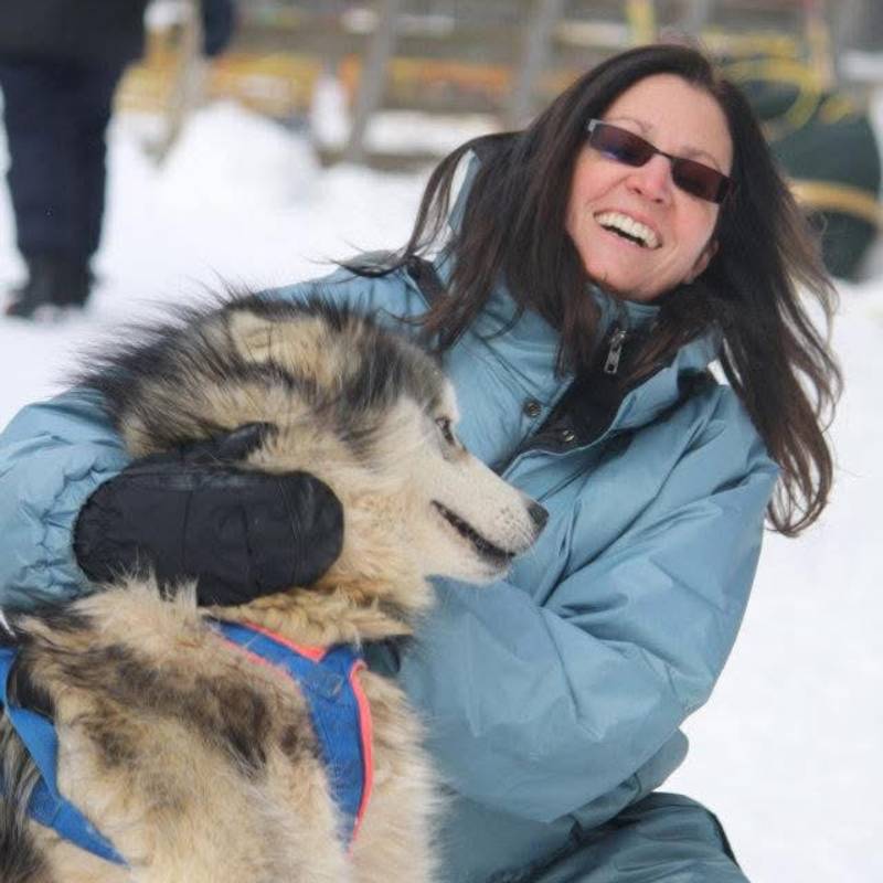 Marie-Claude, in a blue winter jacket, smiles warmly as she holds a large, furry dog outdoors in a snowy setting. Marie-Claude, in a blue winter jacket, smiles warmly as she holds a large, furry dog outdoors in a snowy setting.