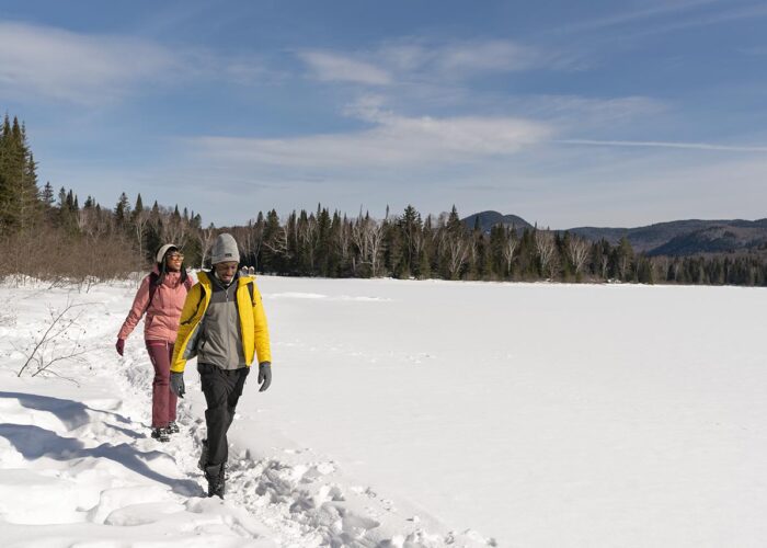 Two people walk along a snowy lakeshore in Mont-Tremblant National Park, surrounded by trees and mountains under a blue sky.