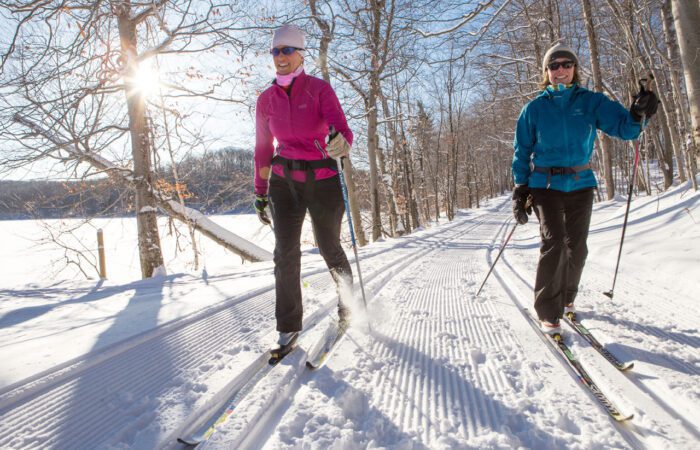 Two people cross-country skiing on a groomed snow trail through a forest in Mont-Tremblant National Park on a sunny winter day.