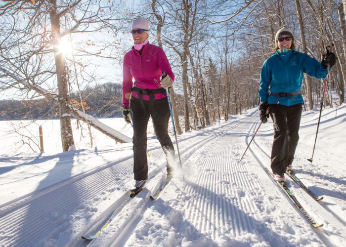 Two people cross-country skiing on a groomed snow trail through a forest in Mont-Tremblant National Park on a sunny winter day.