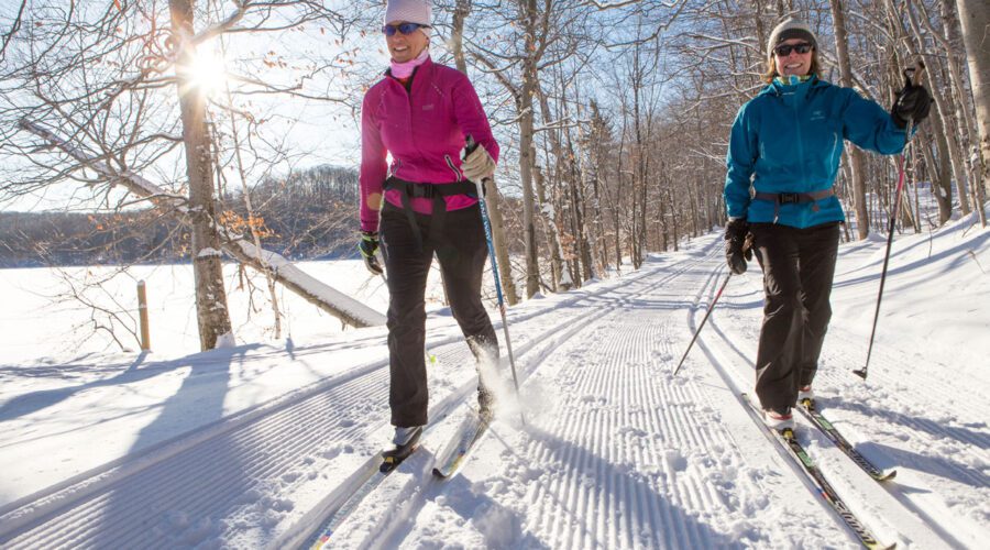 Deux personnes font du ski de fond sur une piste enneigée dans une forêt du parc national du Mont-Tremblant par une journée d'hiver ensoleillée.