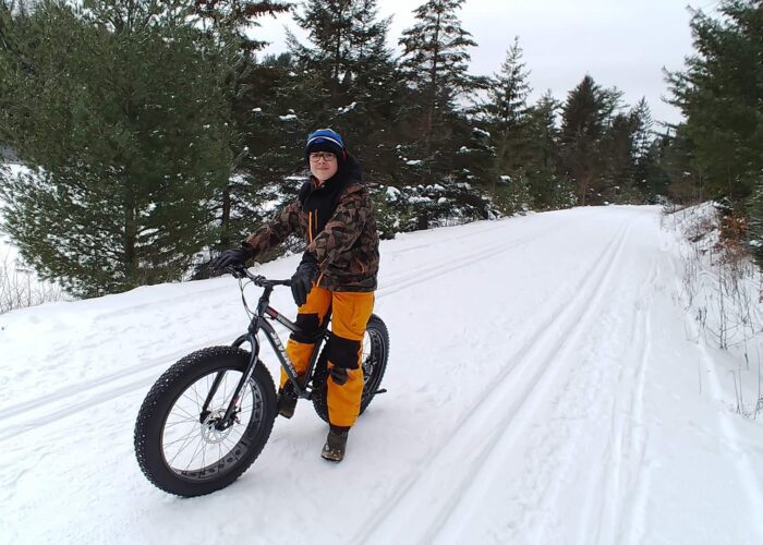 A person in winter clothing stands with a fat-tire bike on a snowy path surrounded by evergreen trees.