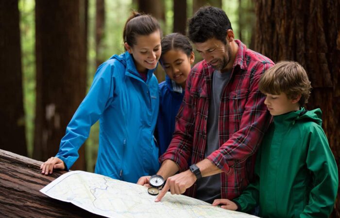 Four people in outdoor clothing examine a map and compass together in a forest setting, standing next to a fallen tree.