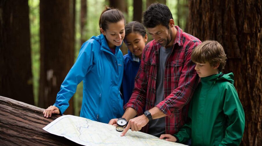 Four people in outdoor clothing examine a map and compass together in a forest setting, standing next to a fallen tree.
