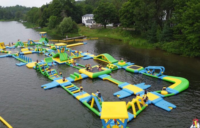 Un grand parcours aquatique gonflable flotte sur un lac, avec des gens qui grimpent, glissent et jouent sur diverses structures colorées. Des arbres et des bâtiments bordent la rive en arrière-plan.