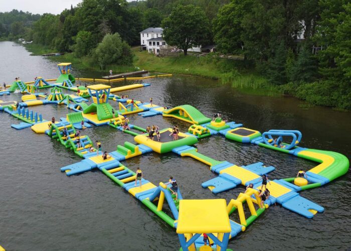 Large inflatable water obstacle course with people climbing, running, and playing on various structures, set on a lake with trees and buildings in the background.