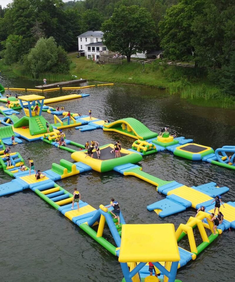 Large inflatable water obstacle course with people climbing, running, and playing on various structures, set on a lake with trees and buildings in the background.