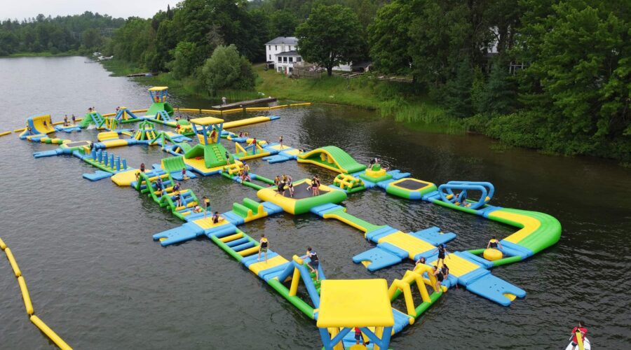 Large inflatable water obstacle course with people climbing, running, and playing on various structures, set on a lake with trees and buildings in the background.