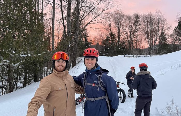 Two people in winter gear and helmets pose on a snowy trail, while two others with bikes stand in the background amid trees at sunset.