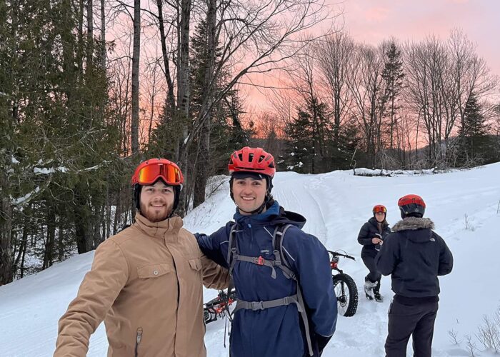 Two people in winter gear and helmets pose on a snowy trail, while two others with bikes stand in the background amid trees at sunset.