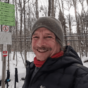 A man in winter clothing smiles outdoors near trail signs in a snowy forest. A man in winter clothing smiles outdoors near trail signs in a snowy forest.