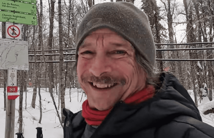 A man in winter clothing smiles outdoors near trail signs in a snowy forest.