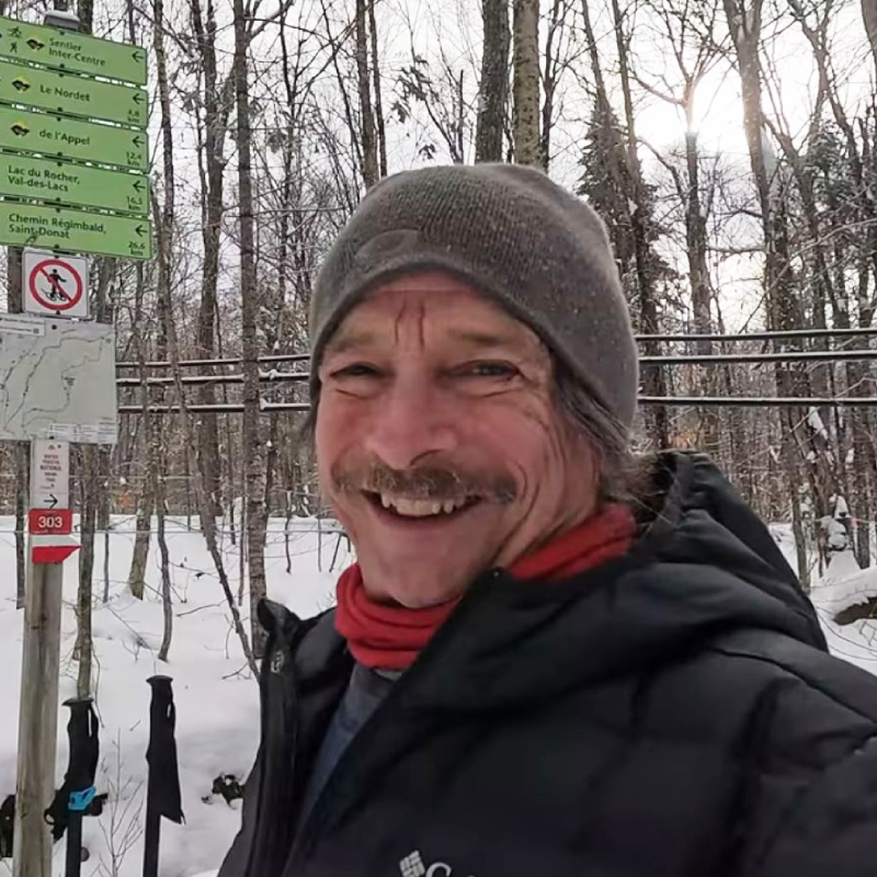 A man in winter clothing smiles outdoors near trail signs in a snowy forest. A man in winter clothing smiles outdoors near trail signs in a snowy forest.
