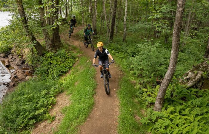Three people ride mountain bikes along a narrow dirt trail through a green forest next to a river.