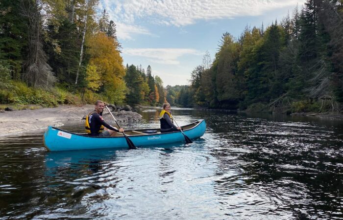 Two people in a blue canoe paddle on a calm river surrounded by trees with autumn foliage under a partly cloudy sky.