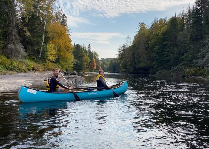 Two people in a blue canoe paddle on a calm river surrounded by trees with autumn foliage under a partly cloudy sky.