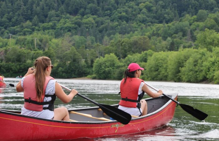 Two people wearing red life vests paddle a red canoe on a calm river surrounded by green trees and forested hills. Two people wearing red life vests paddle a red canoe on a calm river surrounded by green trees and forested hills.