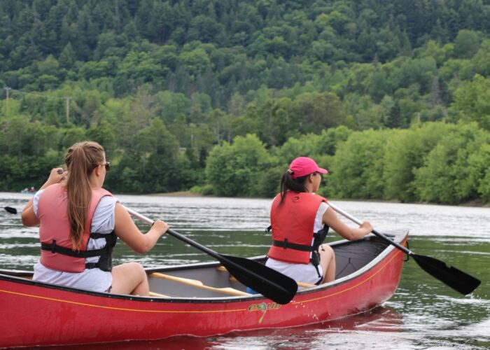 Two people wearing red life vests paddle a red canoe on a calm river surrounded by green trees and forested hills.