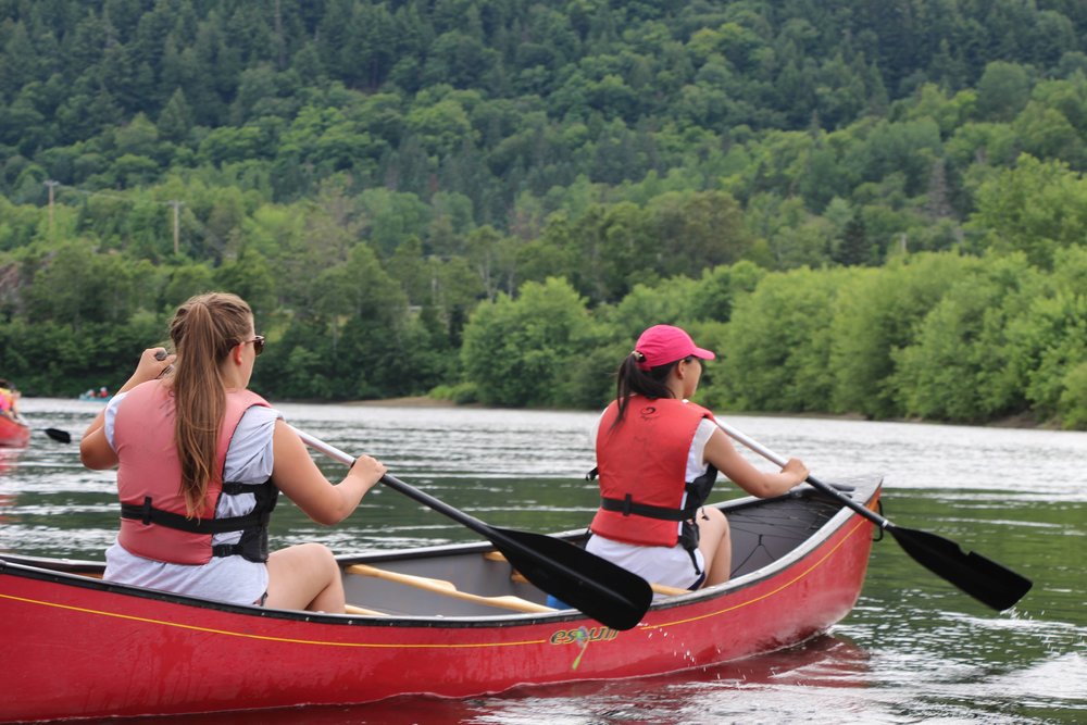 Two people wearing red life vests paddle a red canoe on a calm river surrounded by green trees and forested hills.