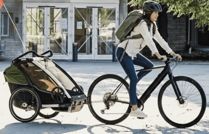 Une personne portant un casque et un sac à dos fait du vélo avec un chariot, tirant une remorque couverte pour enfants le long d’un chemin pavé devant un bâtiment.
