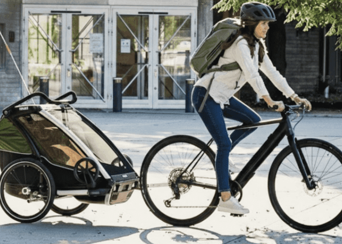 A person wearing a helmet and backpack rides a bike with chariot, pulling a covered child trailer for kids along a paved path in front of a building.