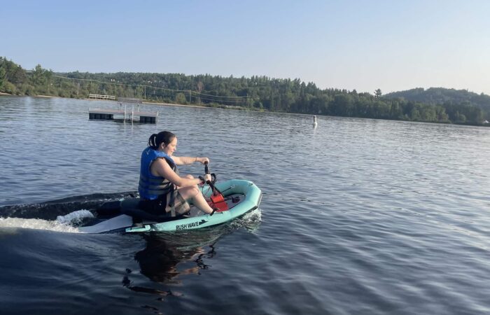 Person wearing a blue life jacket rides a small motorized watercraft on a calm lake with trees and hills in the background.