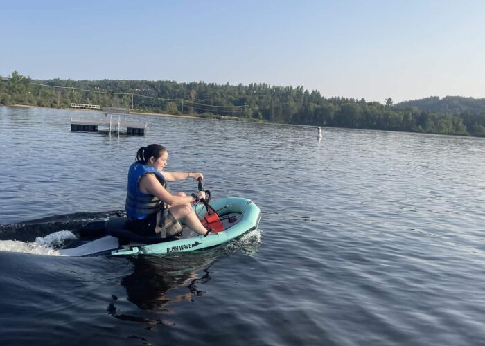 Person wearing a blue life jacket rides a small motorized watercraft on a calm lake with trees and hills in the background.