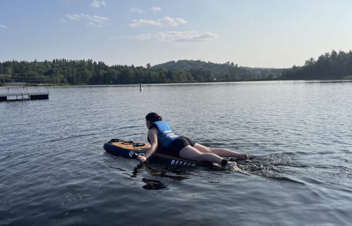 Person wearing a blue life jacket paddles lying down on a paddleboard across a calm lake with trees and a dock in the background.