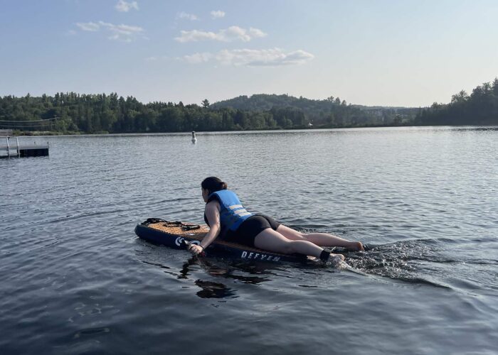 Person wearing a blue life jacket paddles lying down on a paddleboard across a calm lake with trees and a dock in the background.