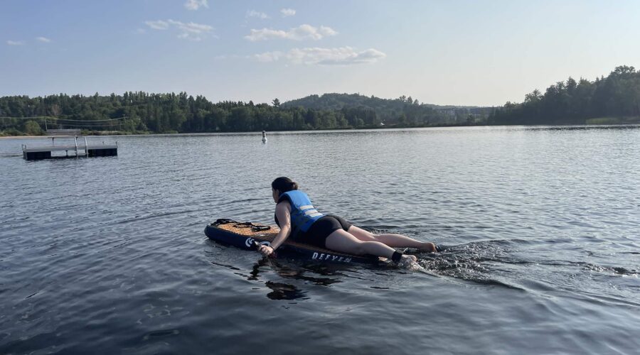 Une personne vêtue d’un gilet de sauvetage bleu pagaie sur un E-bodyboard sur un lac calme, entouré d’arbres sous un ciel clair, profitant des sensations des sports nautiques. Une personne vêtue d'un gilet de sauvetage bleu pagaie sur un E-bodyboard sur un lac calme, entouré d'arbres sous un ciel clair, profitant des sensations des sports nautiques.