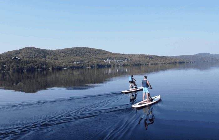 Deux personnes pratiquent des sports nautiques, font du paddleboard sur un lac calme entouré de collines couvertes d'arbres sous un ciel bleu clair.